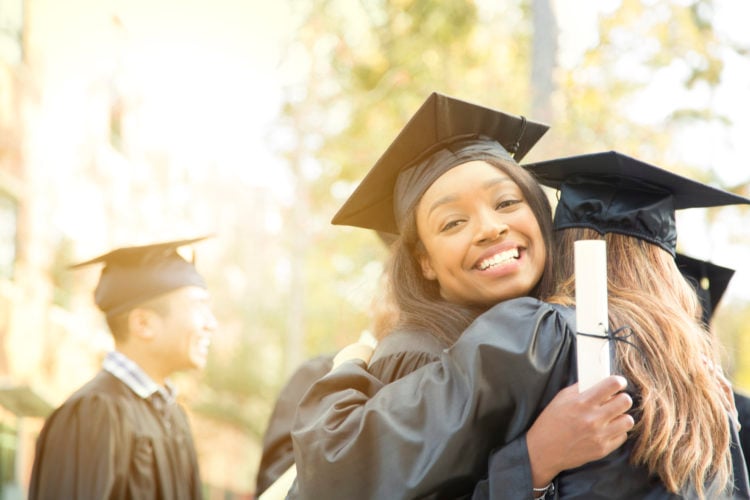 Multi-ethnic group of college student girl friends excitedly give each other big hugs after graduation ceremony.   They stand on campus, hold diplo...