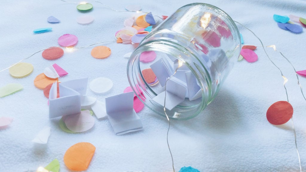 Glass wish jar filled with paper wishes on a table with confetti
