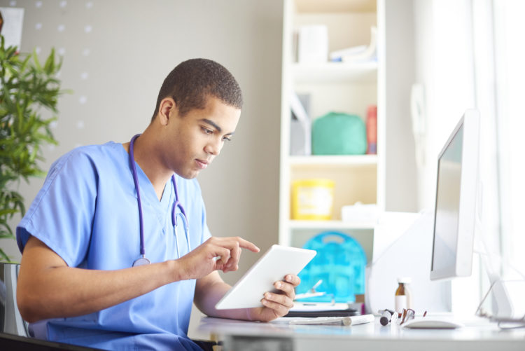 male gp sits at his desk looking at his digital tablet reading through patient notes, emails and looking through patients x-ray's. He is wearing bl...