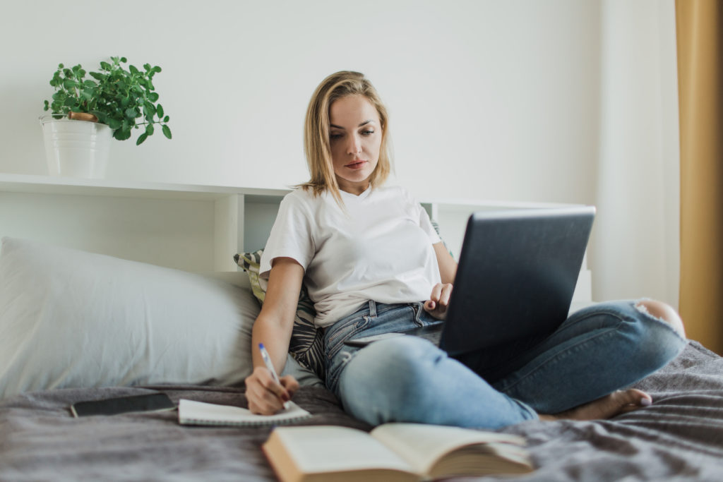 a student girl in a white t shirt and jeans is sitting on the bed studying writing reading