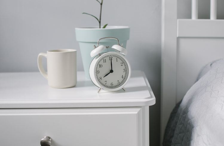 White alarm clock, beige cup with coffee, tea, house plant on bedsite in bedroom. Morning concept. Scandinavian light interior