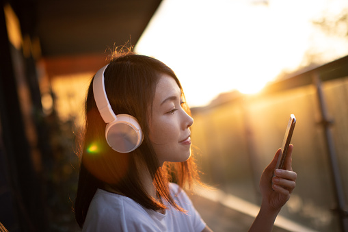Beautiful young Asian woman listening to podcast with wireless headphone and smartphone, relaxing on the balcony at sunset.