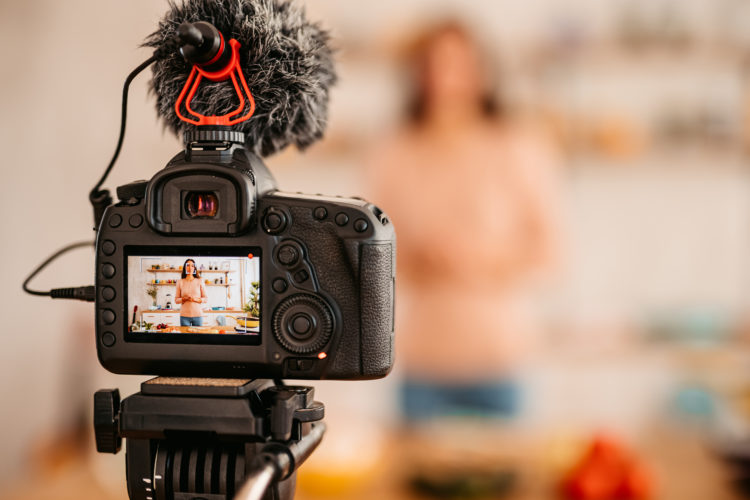 Cheerful young woman blogger recording live tutorial video, about healthy vegetarian eating on camera of in kitchen.