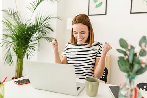 Cheerful young woman celebrating her achievement while reading good news on laptop from home office - Smiling female expressing excitement working ...