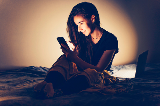 Young woman is using mobile phone at night sitting on bed