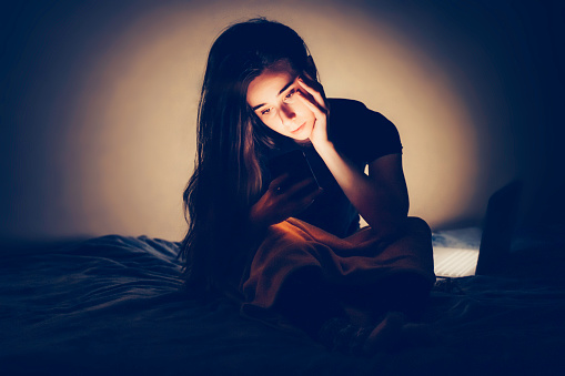 Young woman is using mobile phone at night sitting on bed. She is texting her friend via social media or messenger, smiling with joy. Internet addi...