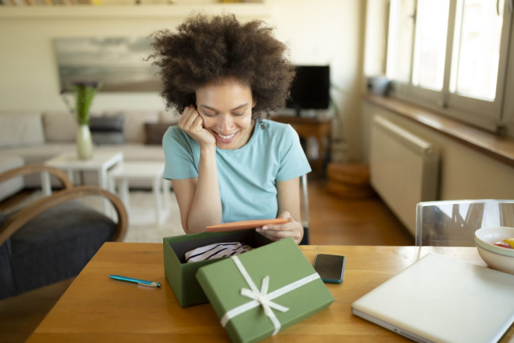 Cheerful African American woman with a beautiful smile, opening a gift at home, feeling happy and reading a letter she received with it