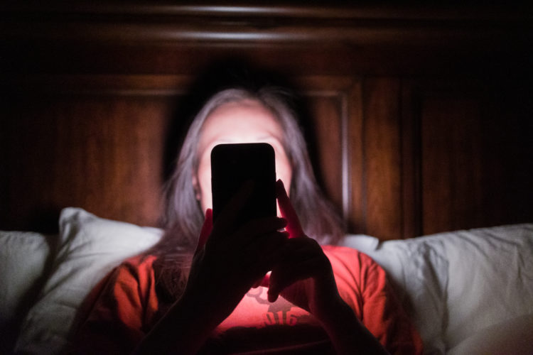 Woman laying in bed and checking updates on smartphone. Face and bed illuminated by light emitted from display. Screen addiction and modern technol...