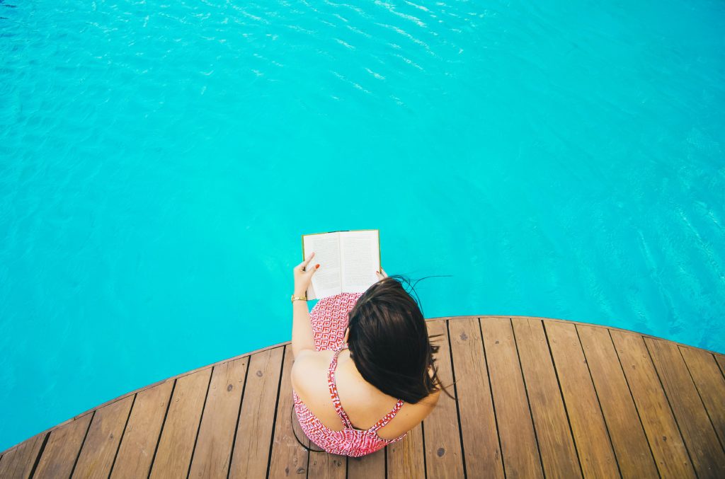woman sitting on poolside dock while riding book