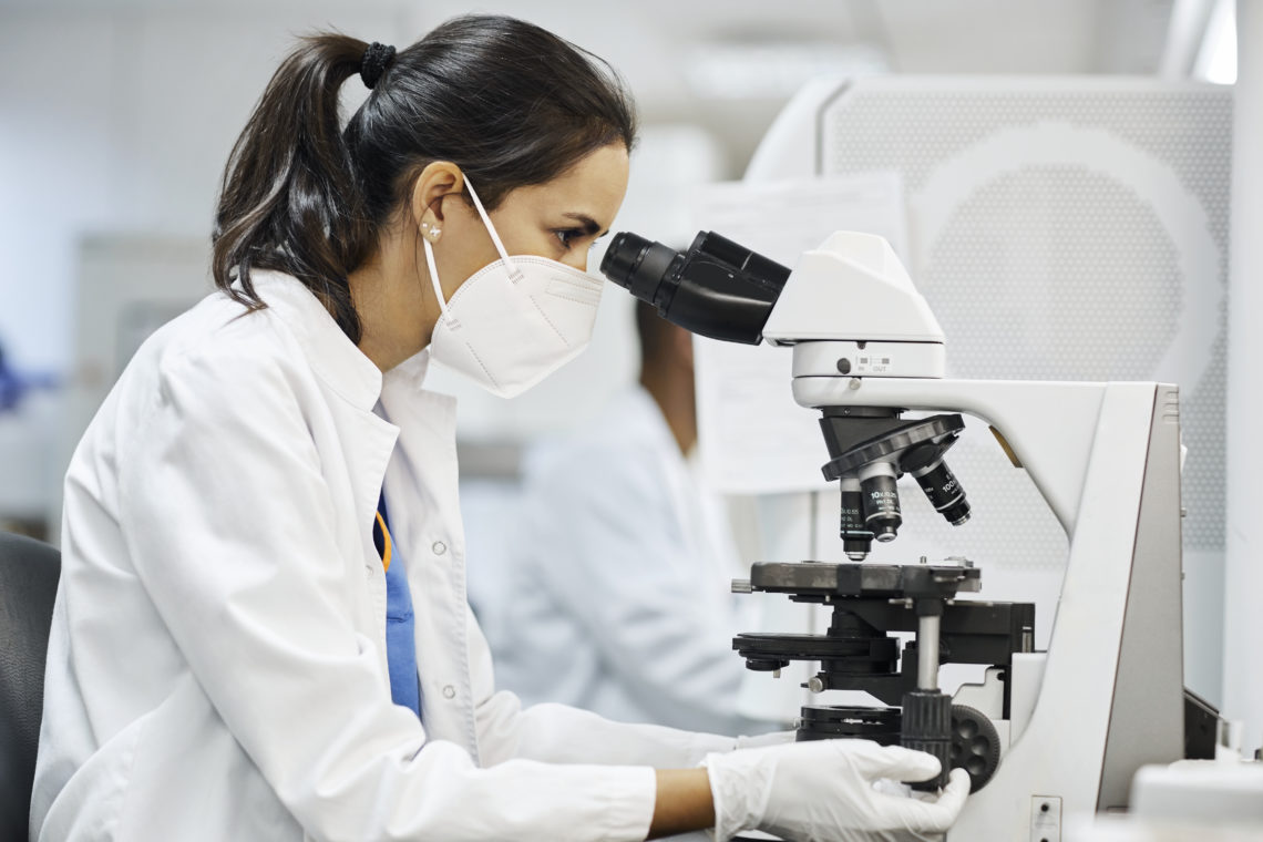 Side view of female doctor using microscope during research in laboratory. Healthcare professional is working in hospital during COVID-19. She is w...
