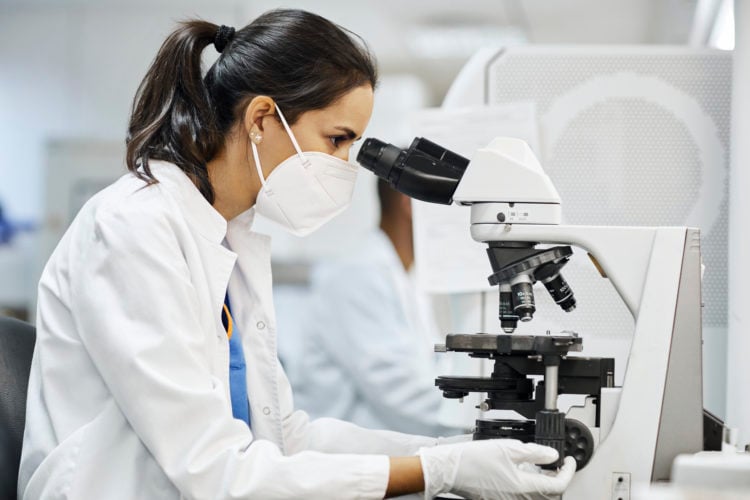 Side view of female doctor using microscope during research in laboratory. Healthcare professional is working in hospital during COVID-19. She is w...