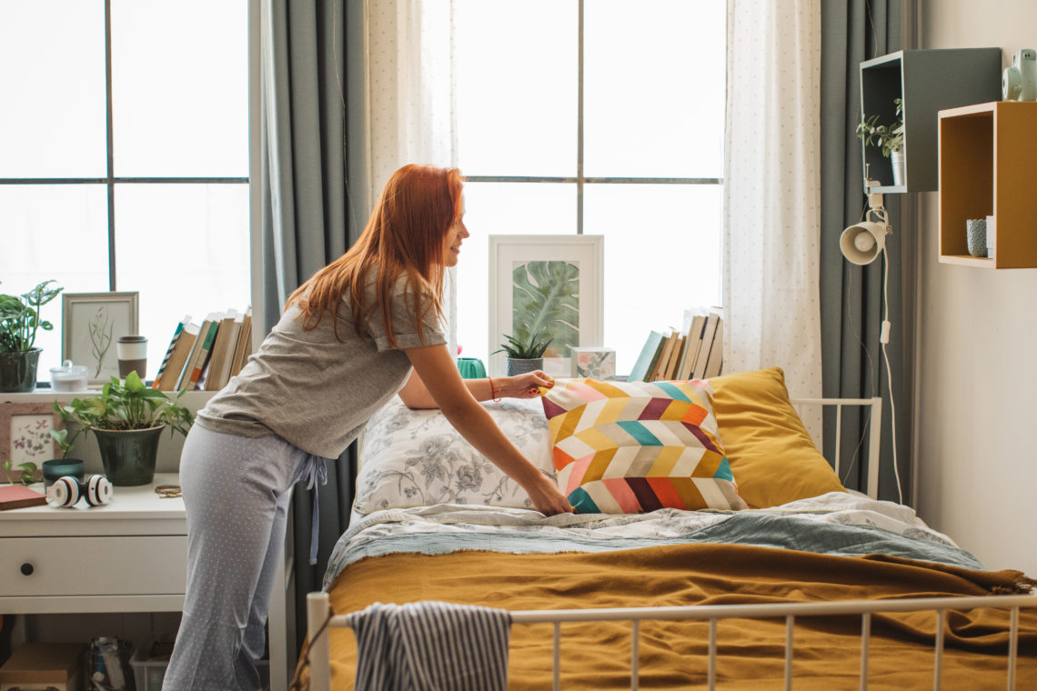 Young woman at dorm room. It is morning and she is making bed while listening music on headphones.