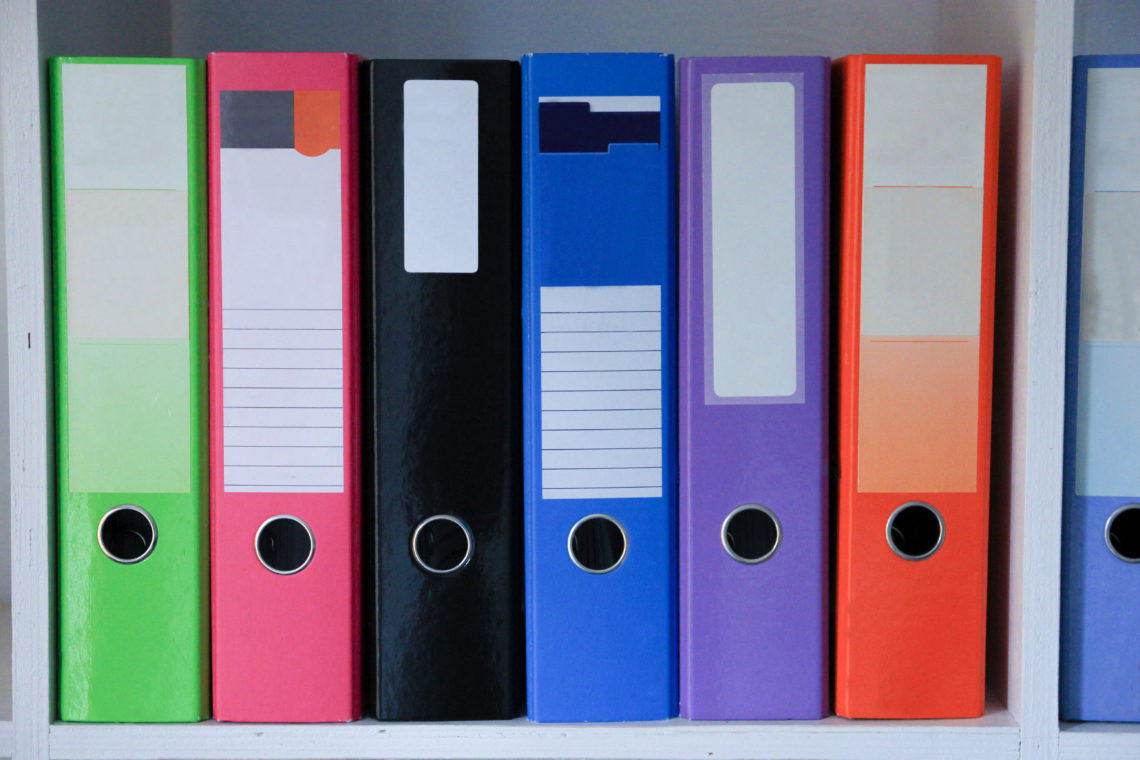 Row of brightly coloured A4 lever arch files on a white shelf used for storing documents.