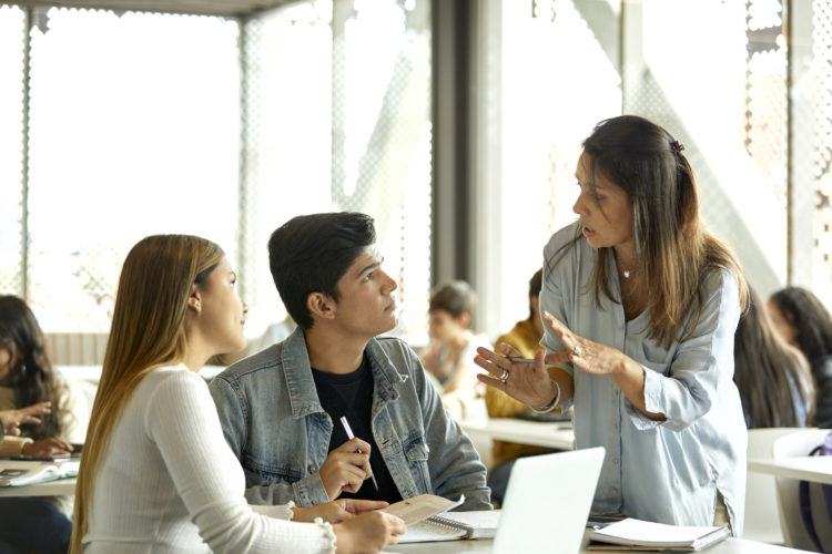 Mature teacher explaining students in classroom. Male and female are listening to professor in university. They are sitting at table.