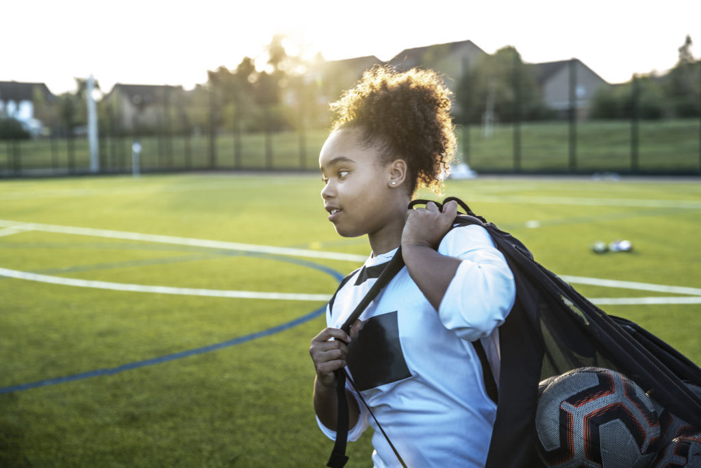 UK, Rear view of female soccer player carrying bag with balls in field