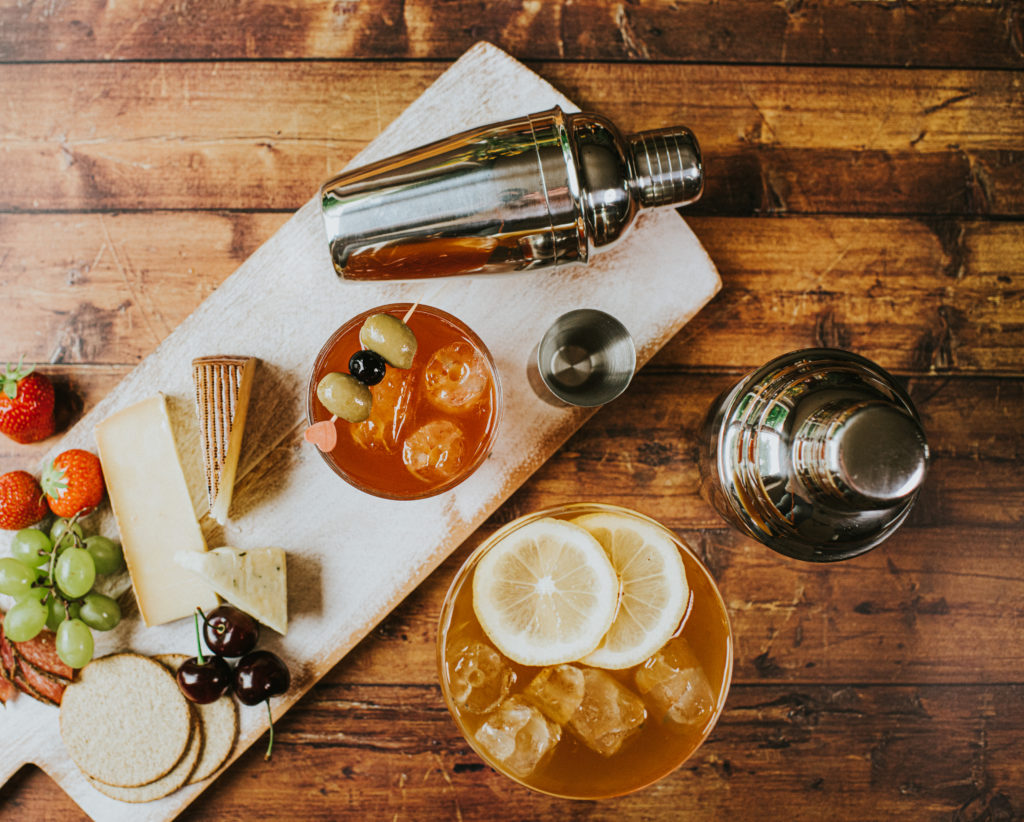 Top-down image of a delicious, simple cheese board and a cocktails on a wooden table beside cocktail shakers