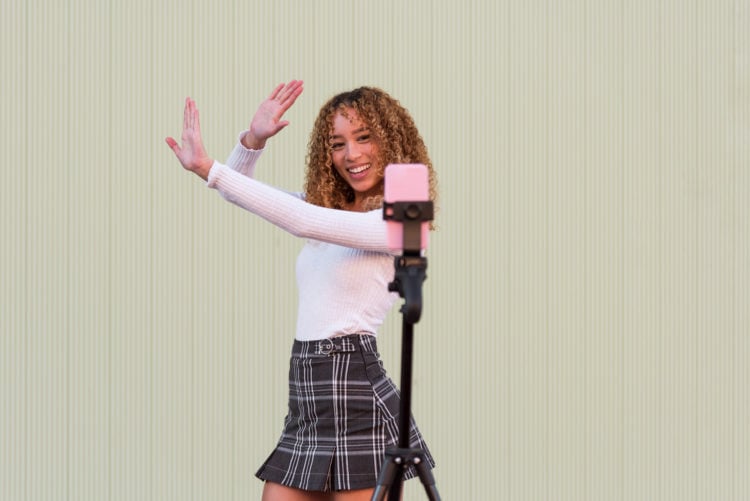 Positive Hispanic female blogger in stylish wear with curly hair dancing and recording video for Tik Tok on cellphone while standing near wall on s...
