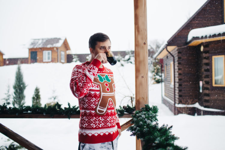 Young man in a Christmas sweater warms up with hot drink on the terrace of a modern house during the winter holidays.