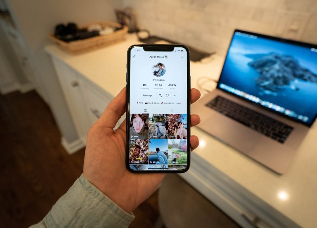person holding phone displaying tiktok profile with desk and computer in background