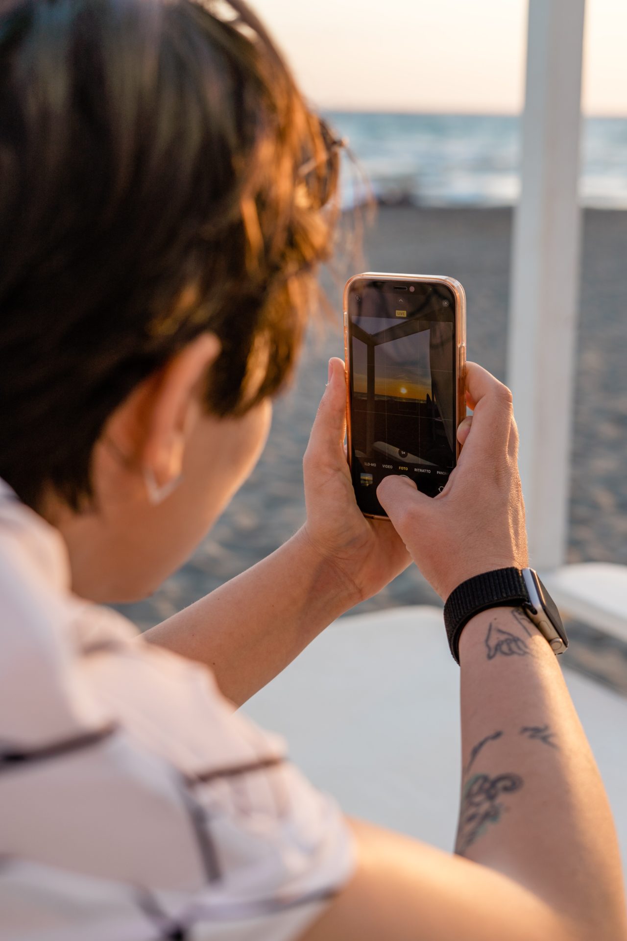 Tattooed girl photographing a sunset at the beach on her iPhone in Rome, Italy