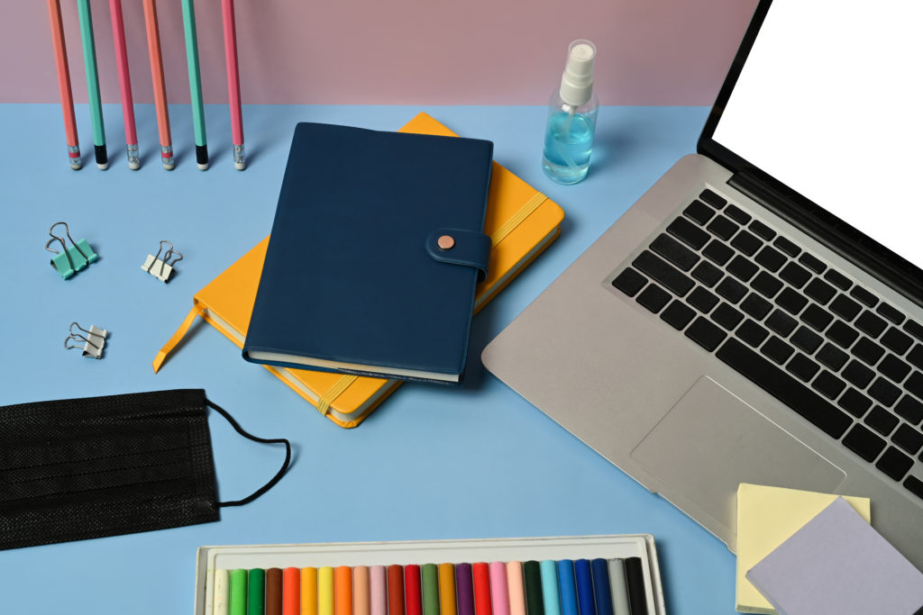 Laptop with white screen, face mask, different stationery on light blue blackboard.