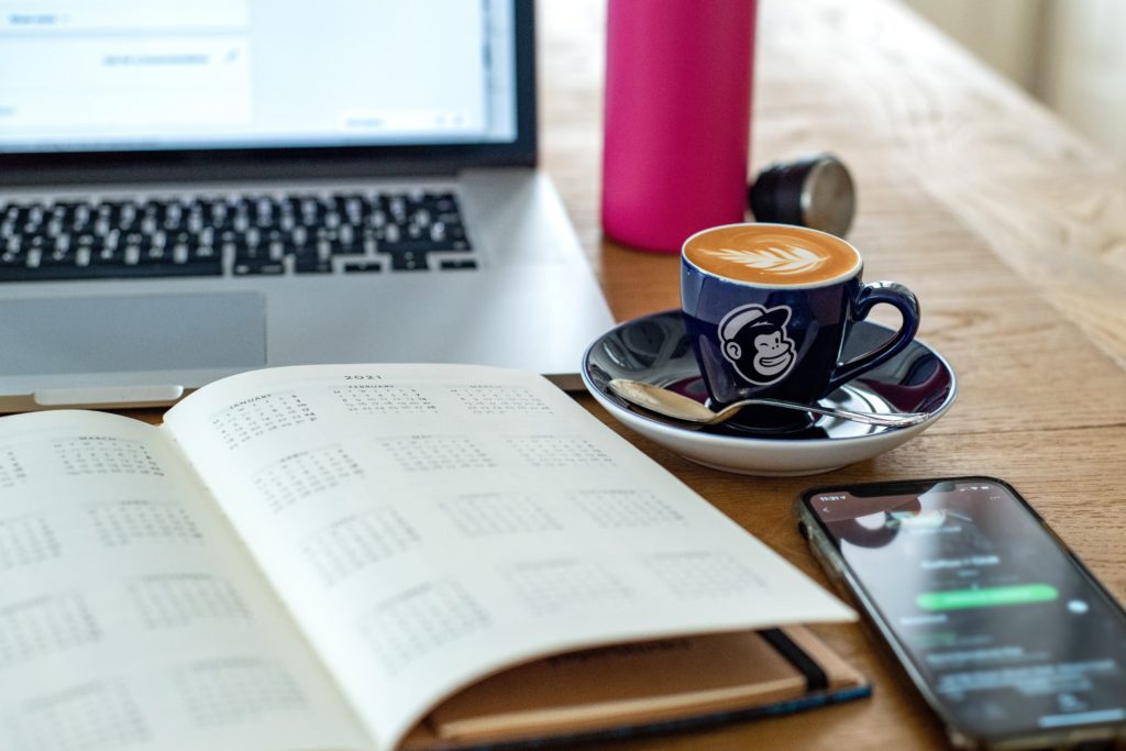 Spotify on a phone next to a diary, a coffee and a computer on a table.