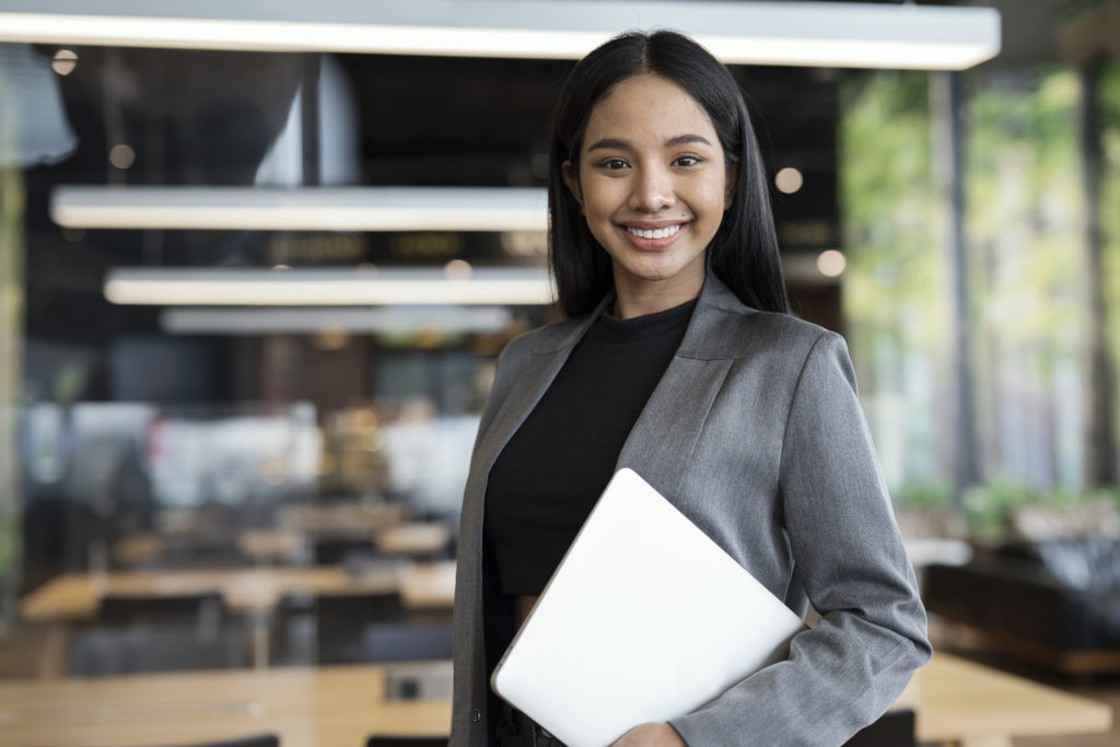 Smart People with Technologies to Boost Business Success. Portrait of Confident Young Female Entrepreneur standing while holding a laptop in a modern business office.