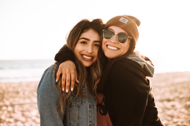 woman hugging other woman while smiling at beach facing the camera