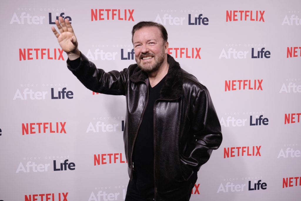 Ricky Gervais wearing black leather jacket and trousers with one arm outstretched waving at Afterlife premier