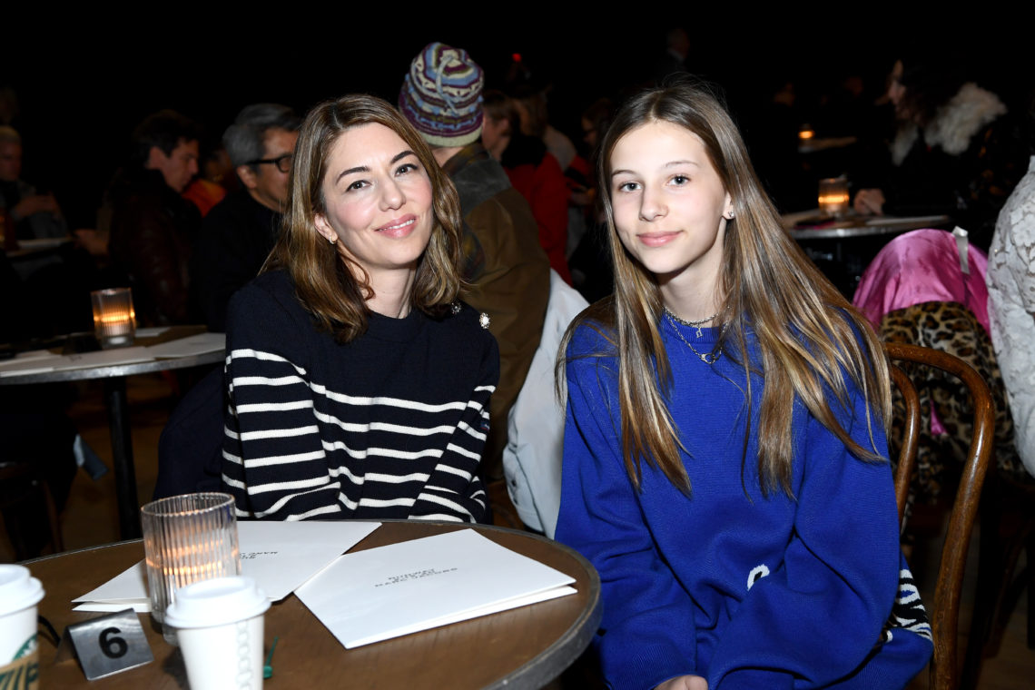 Sofia Coppola in black and white stiped jumper sat at wooden table next to Romy Mars in blue jumper. Both smile at camera with paper and drinks on table