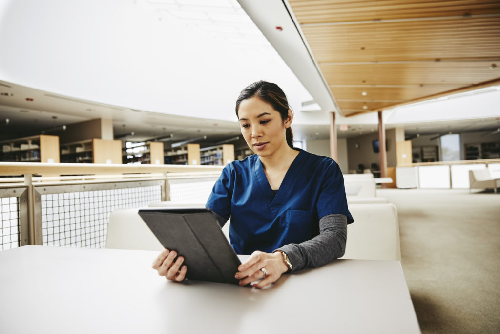 Female medical student in blue scrubs using tablet sat at a white desk in library