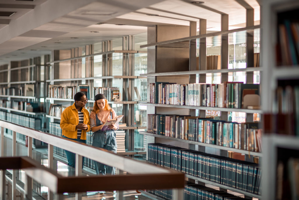 two female students stood together walking through university library holding books