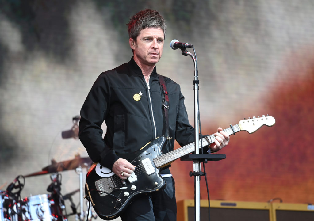 Noel Gallagher on stage playing black guitar in front of microphone in black jacket and jeans