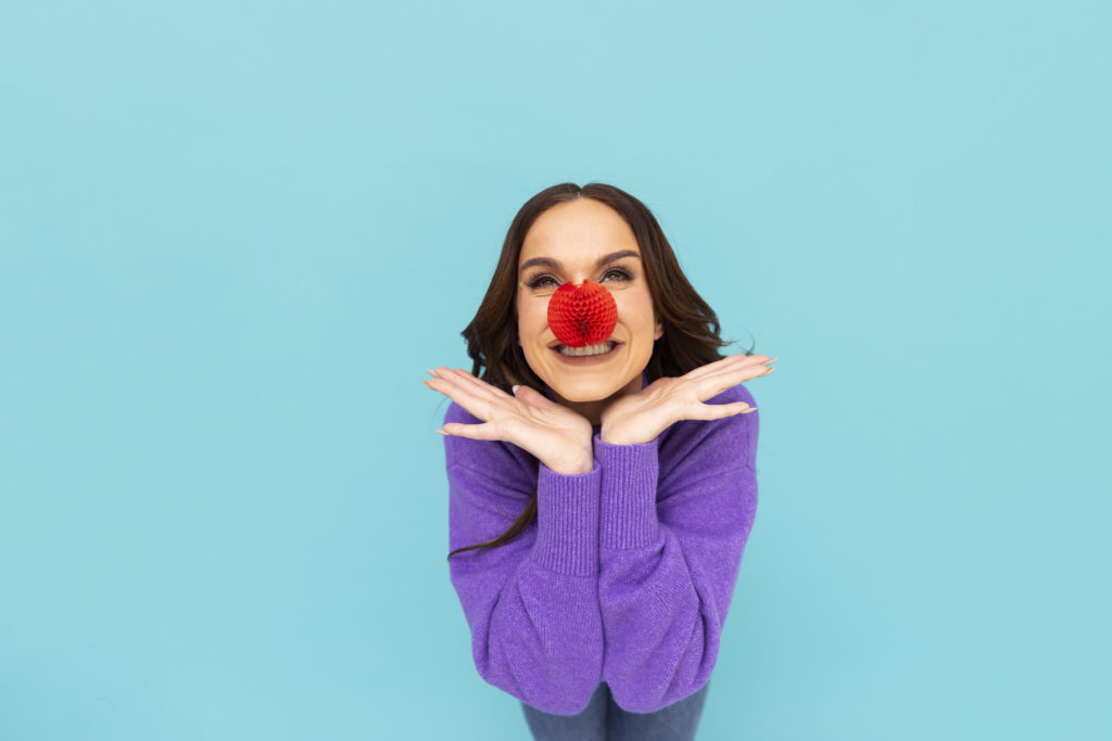 Vicky Pattinson wearing purple jumper and red nose bent towards the camera with chin hovering above hands positioned in a vee