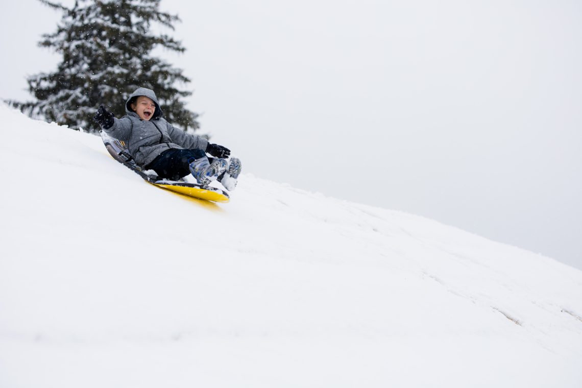 boy in grey jacket riding on yellow sledge down snowy hill