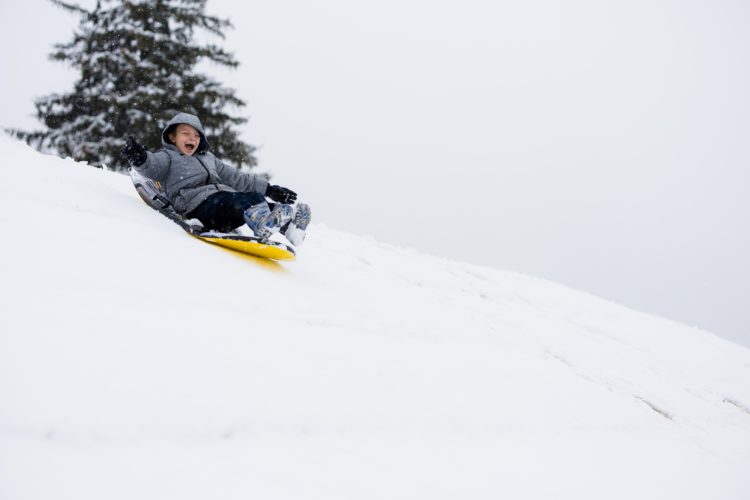 boy in grey jacket riding on yellow sledge down snowy hill