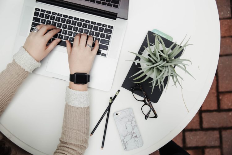 person in a beige jumper using laptop computer beside aloe vera, pencils and a phone.