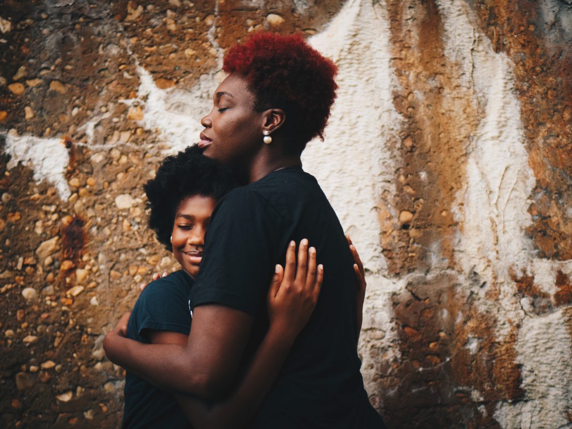 a mum hugging a daughter in front of stone wall. Bith are wearing black tshirts and smiling