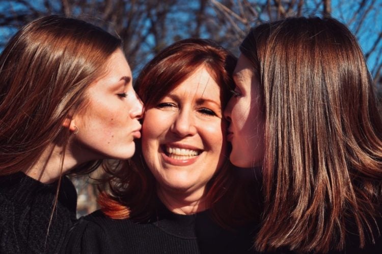 three women in black tshirts. Two are kissing the middile one on the cheek as she smiles