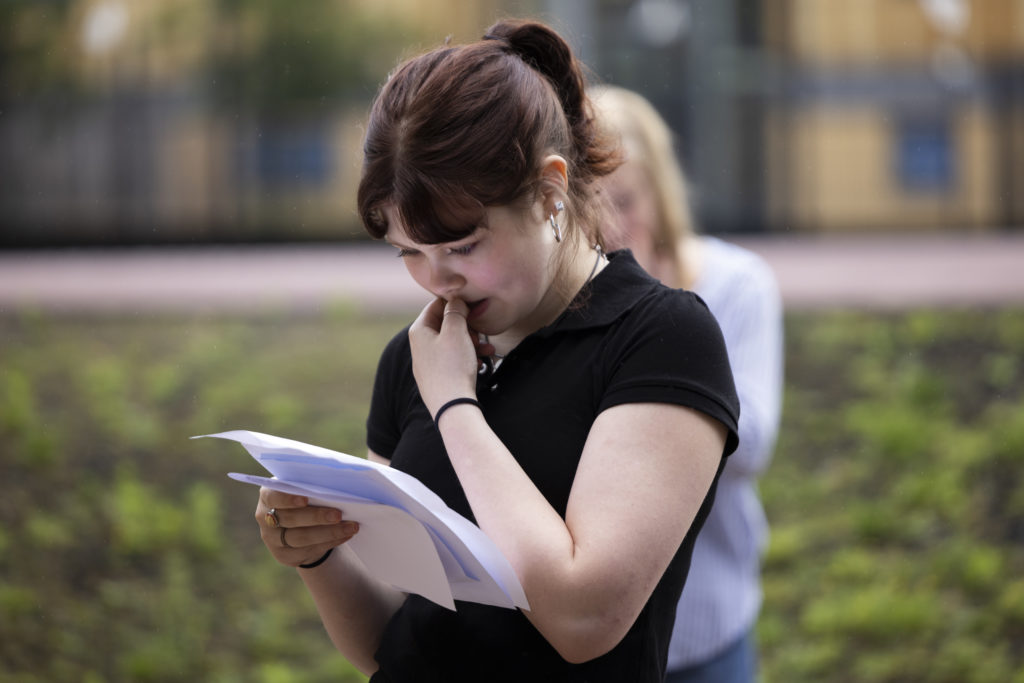 Student in black tshirt and brown hair in ponytail looking worried with hand to mouth reading GCSE results