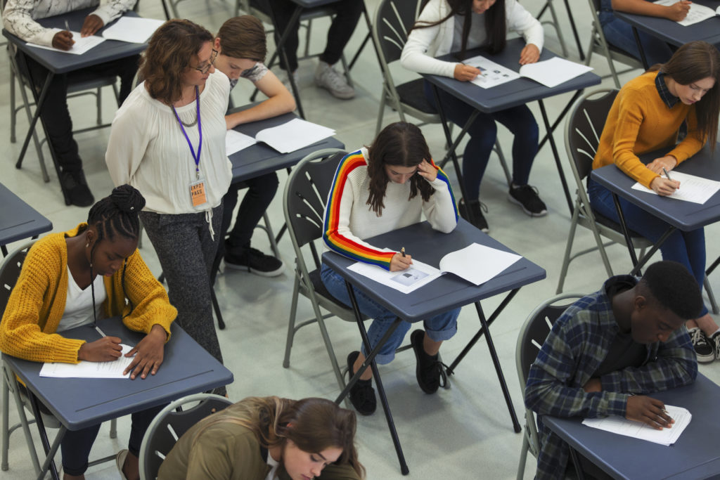 a group of students take an exam sitting at separate blue tables as an invigilator walks past