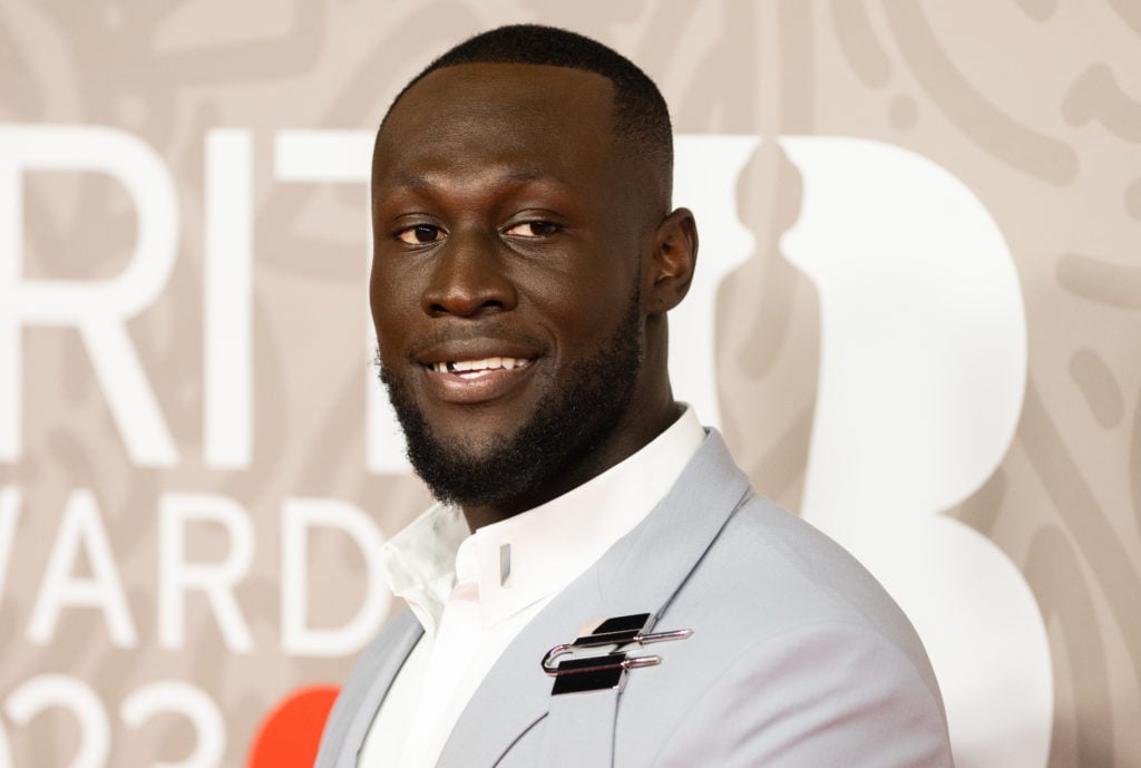 Stormzy stoof side on facing camera in light grey suit with white shirt at Brit Awards 2023