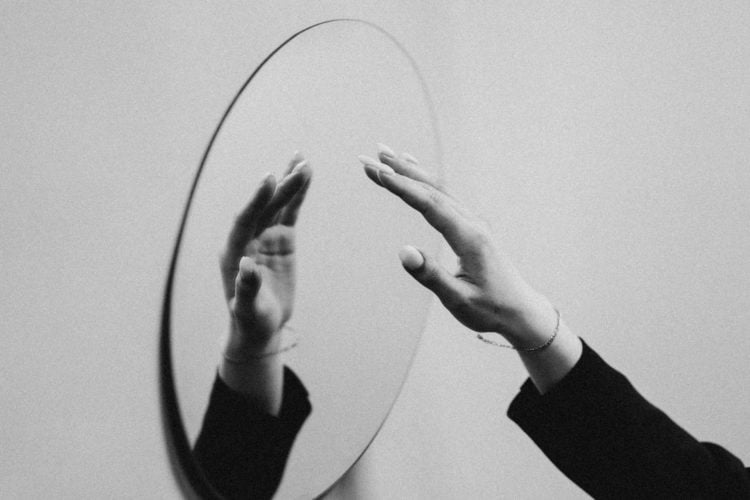 b&w photo of person in black long sleeve shirt reaching towards oval mirror on wall