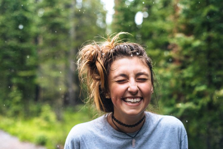 woman outdoors smiling with whole face. Her hair is in messy bun and she's wearing a grey jumper