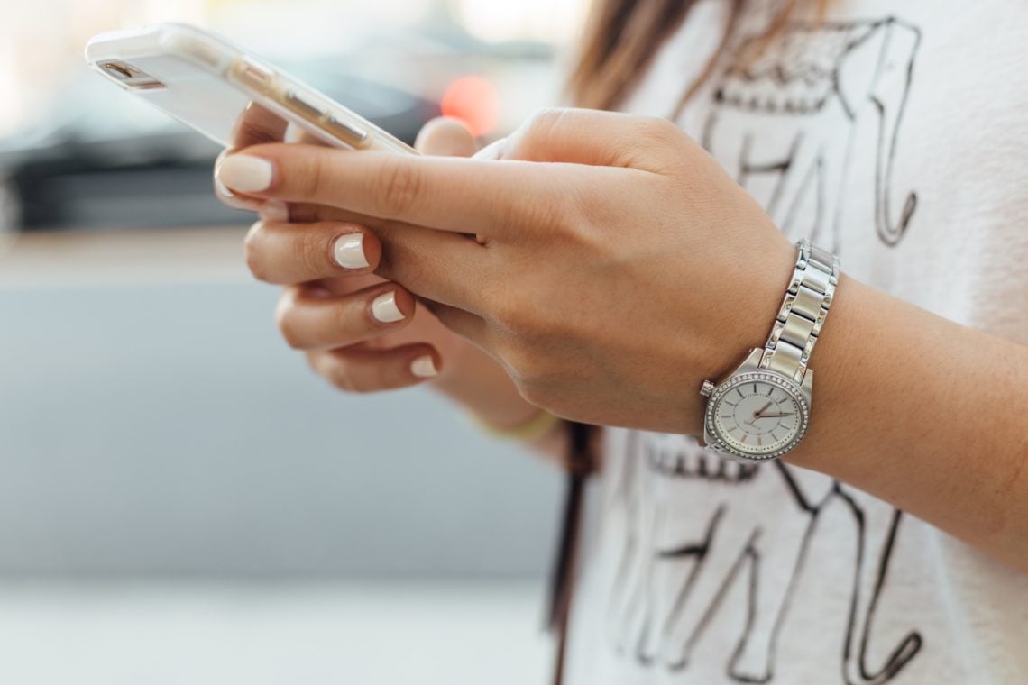 close up of woman holding iPhone during daytime. She has white painted nails, a silver watch and a white tshirt