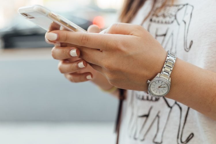 close up of woman holding iPhone during daytime. She has white painted nails, a silver watch and a white tshirt