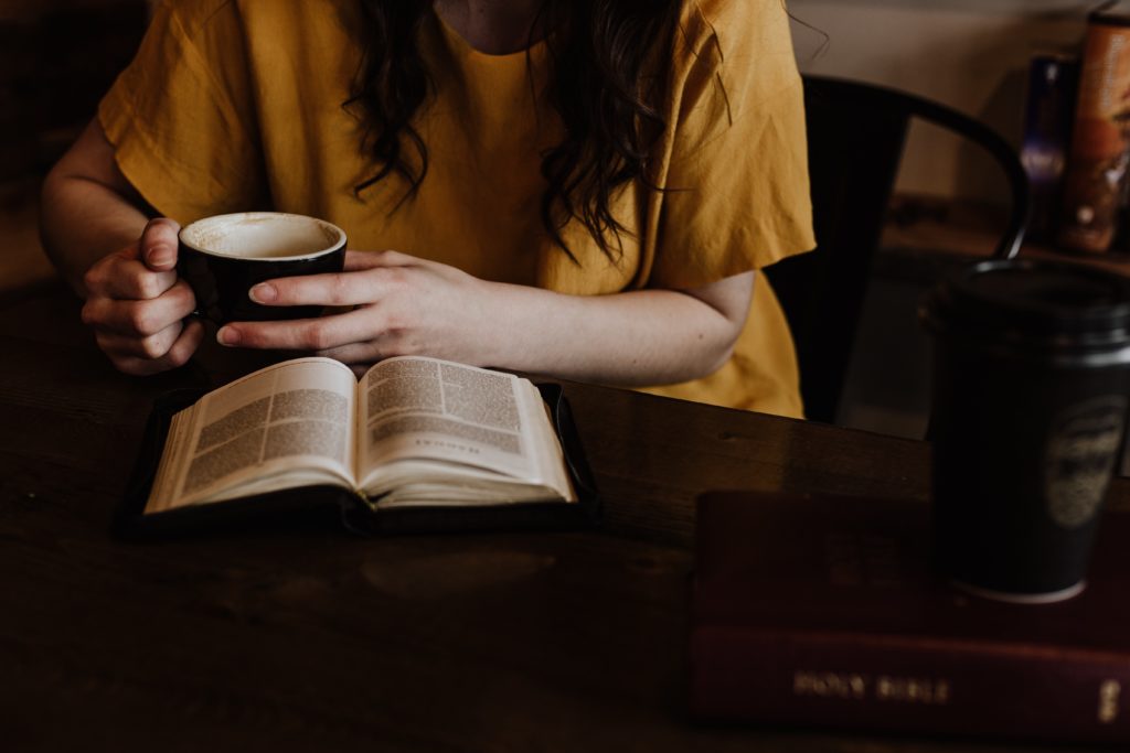 woman in yellow tshirt holding black mug, sitting at table with book open.