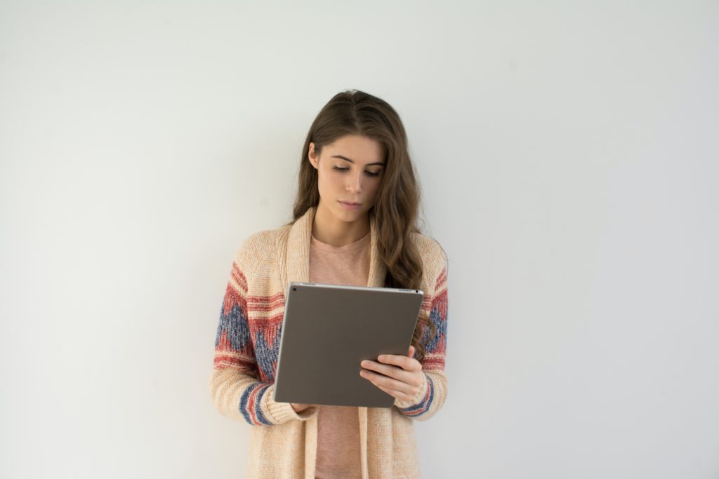 woman in beige, gray, and red sweater holding silver tablet computer in front of white wall