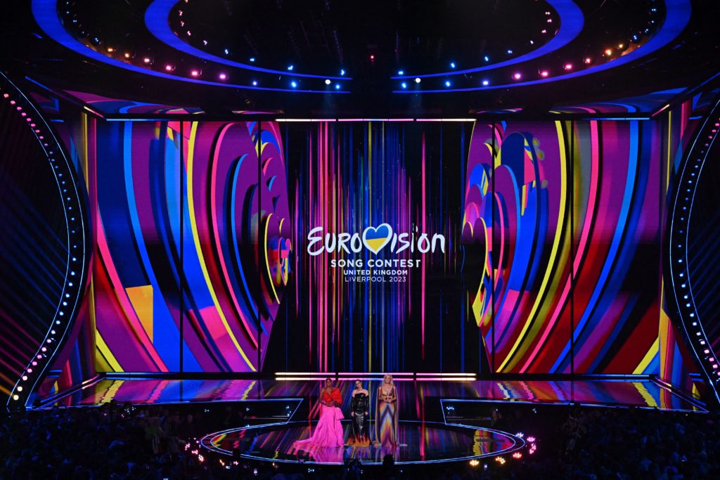 Wide shot of Eurovision stage in liverpool with hosts Alesha Dixon in pink and red dress, Julia Sanina in black dress and Hannah Waddingham in rainbow dress