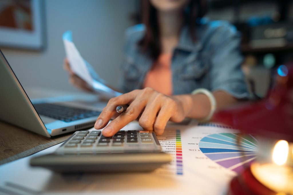 close up of female hang tapping a calculater as she hold paper in front of laptop in blurred background. She wears an orange top with a denim jacket